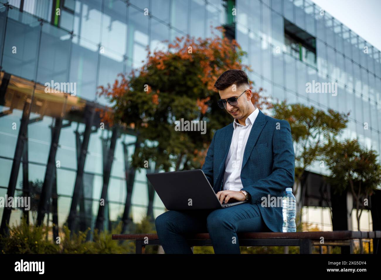 Ein Mann sitzt auf einer Bank in einem öffentlichen Bereich, vertieft in seine Arbeit auf einem Laptop. Er scheint auf dem Bildschirm konzentriert zu sein. Stockfoto
