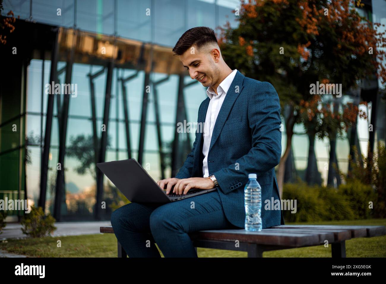 Ein professioneller Mann in einem formellen Anzug sitzt auf einer Bank, während er auf seinem Laptop schreibt. Sein zielgerichtetes Verhalten deutet darauf hin, dass er wahrscheinlich an wichtig arbeitet Stockfoto
