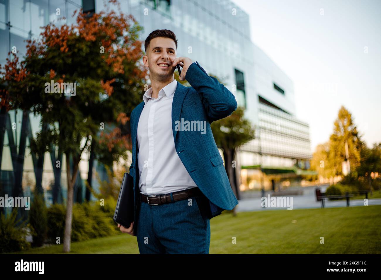 Ein junger Mann in blauem Anzug und weißem Hemd steht vor einem Bürogebäude, hält einen Laptop und spricht mit seinem Handy. Stockfoto