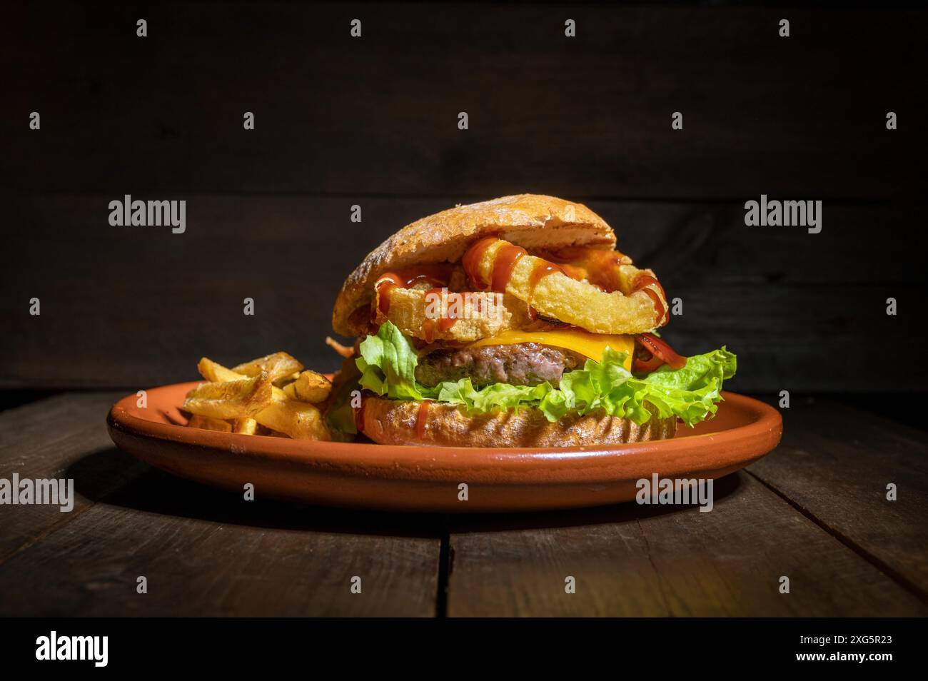 Premium-Burger mit Zwiebelringen, Käse und Grillsauce, serviert mit pommes Frites auf einem rustikalen Teller. Hochwertige Fotografie Stockfoto