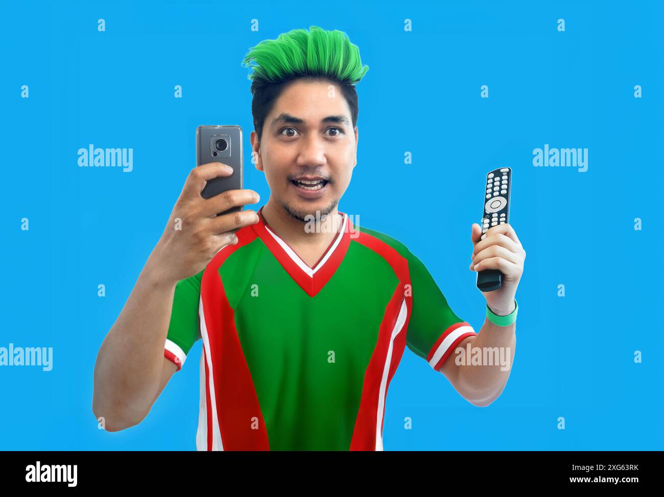 Asiatischer Mann im Fußball Protugal Trikot halten Handy und tv-Fernbedienung wie wie oder Siegerwette Spiel auf isoliertem Hintergrund Stockfoto