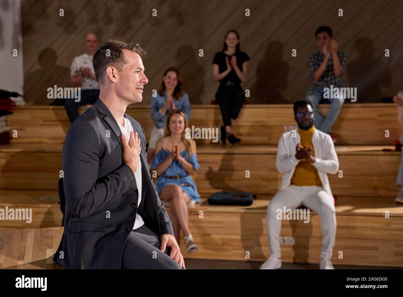 Couch im formellen Anzug, der Dankbarkeit für die Anerkennung durch die Zuhörer ausdrückt, Hände auf der Brust hält und fröhlich lächelt. Gruppe von Erwachsenen, die die Hände klatschen Stockfoto