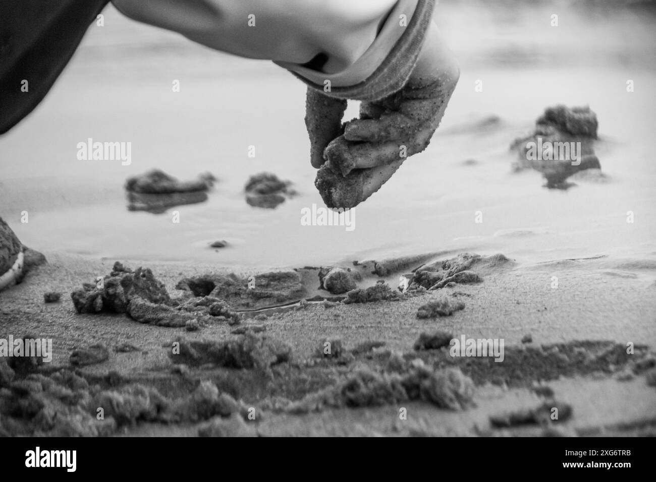 Ein schwarzweißes Nahfoto einer Kinderhand, die mit nassem Sand am Strand spielt. Stockfoto