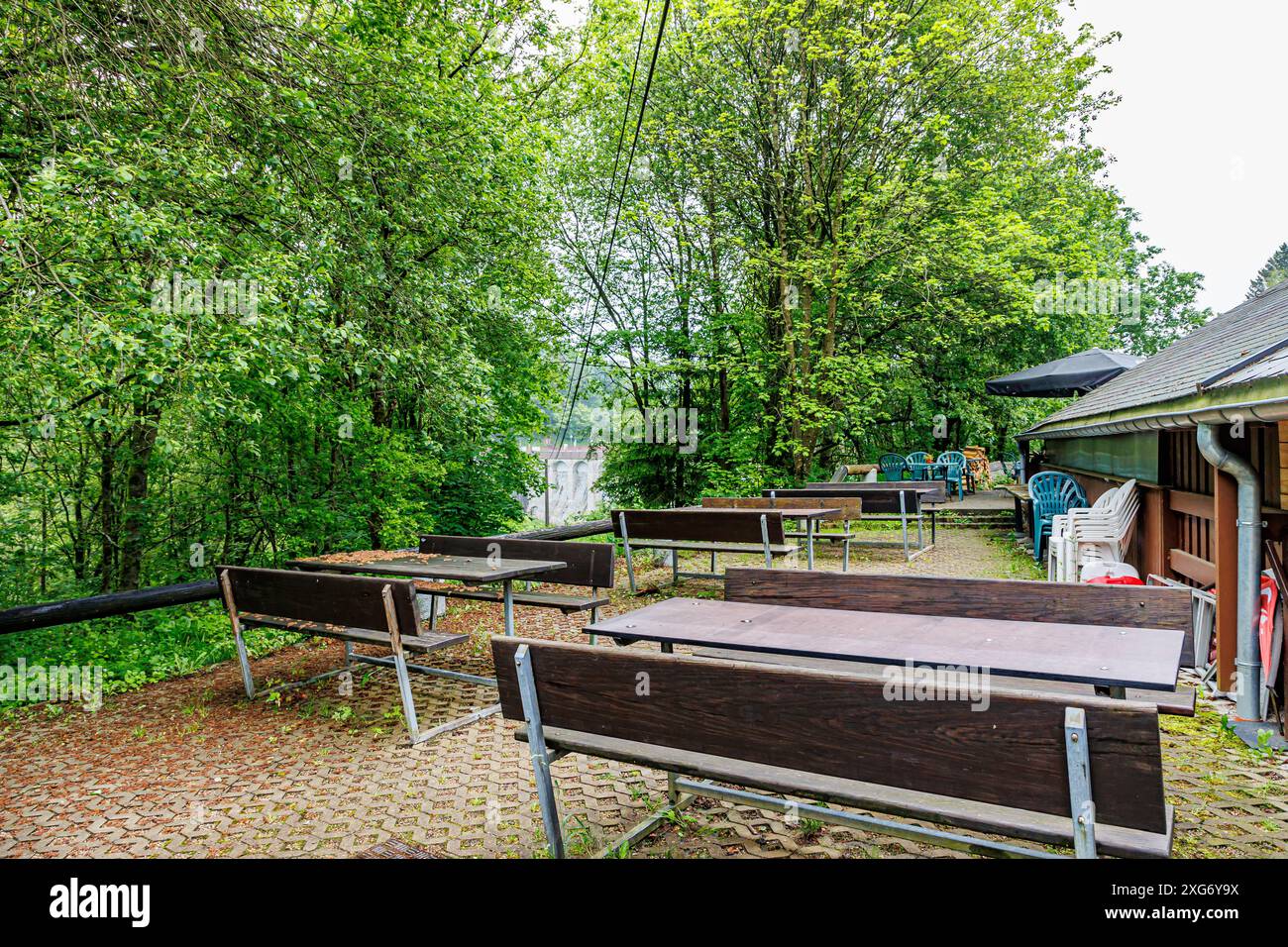 Rustikale Hinterterrasse eines Landrestaurants, Holztische und -Bänke, Plastikstühle neben der Wand, Laubbäume mit grünem Laub im Hintergras Stockfoto