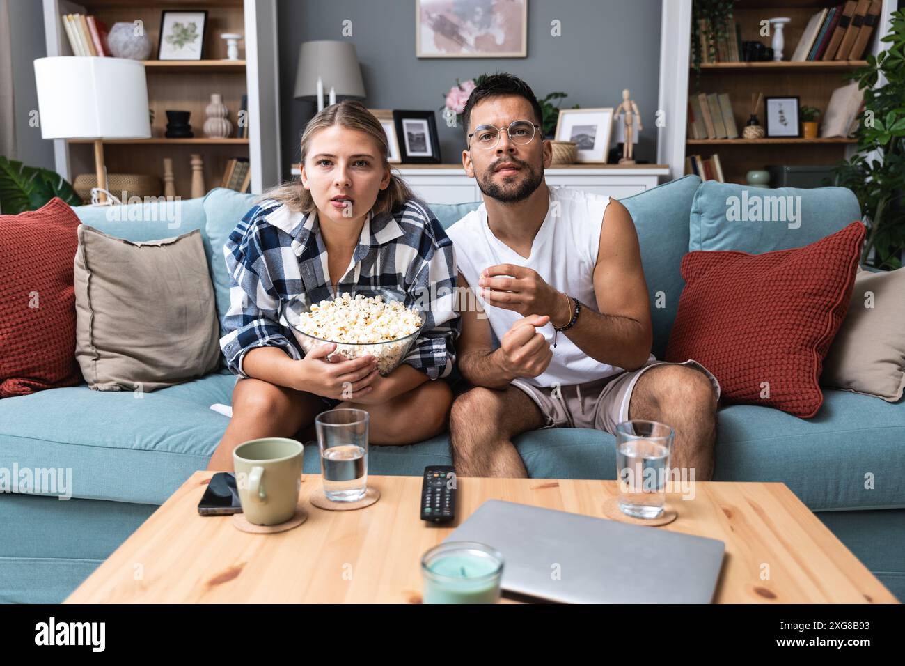 Ein junges Paar, Mann und Frau, sitzt zu Hause auf dem Sofa, isst Popcorn und schaut schaurige Horrorfilme an oder sieht sich dramatische Sportaktivitäten oder Dramen an Stockfoto Ein junges Paar, Mann und Frau, sitzt zu Hause auf dem Sofa, isst Popcorn und schaut schaurige Horrorfilme an oder sieht sich dramatische Sportaktivitäten oder Dramen an Stockfoto