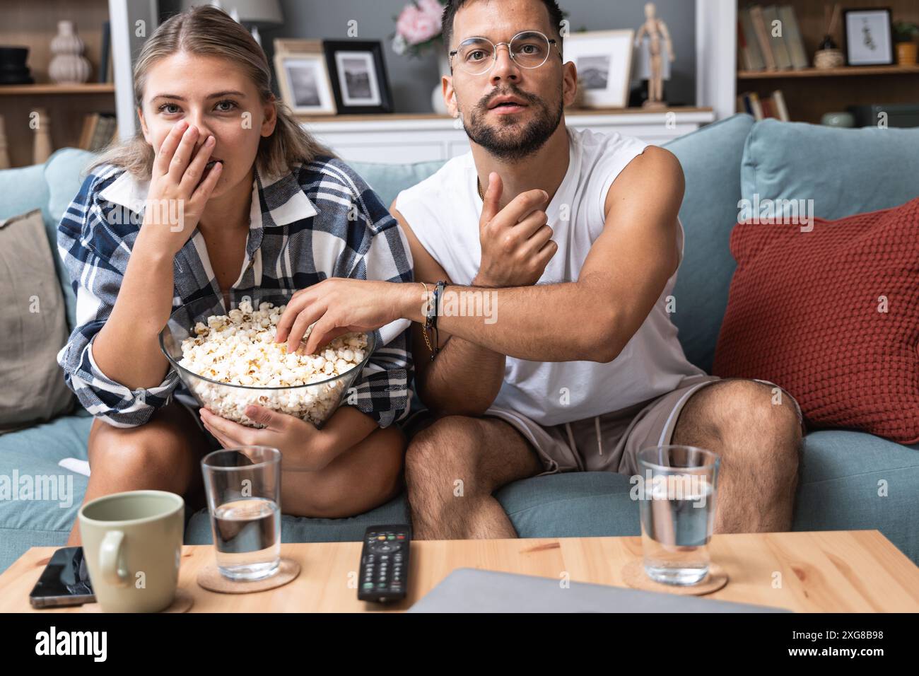 Ein junges Paar, Mann und Frau, sitzt zu Hause auf dem Sofa, isst Popcorn und schaut schaurige Horrorfilme an oder sieht sich dramatische Sportaktivitäten oder Dramen an Stockfoto Ein junges Paar, Mann und Frau, sitzt zu Hause auf dem Sofa, isst Popcorn und schaut schaurige Horrorfilme an oder sieht sich dramatische Sportaktivitäten oder Dramen an Stockfoto