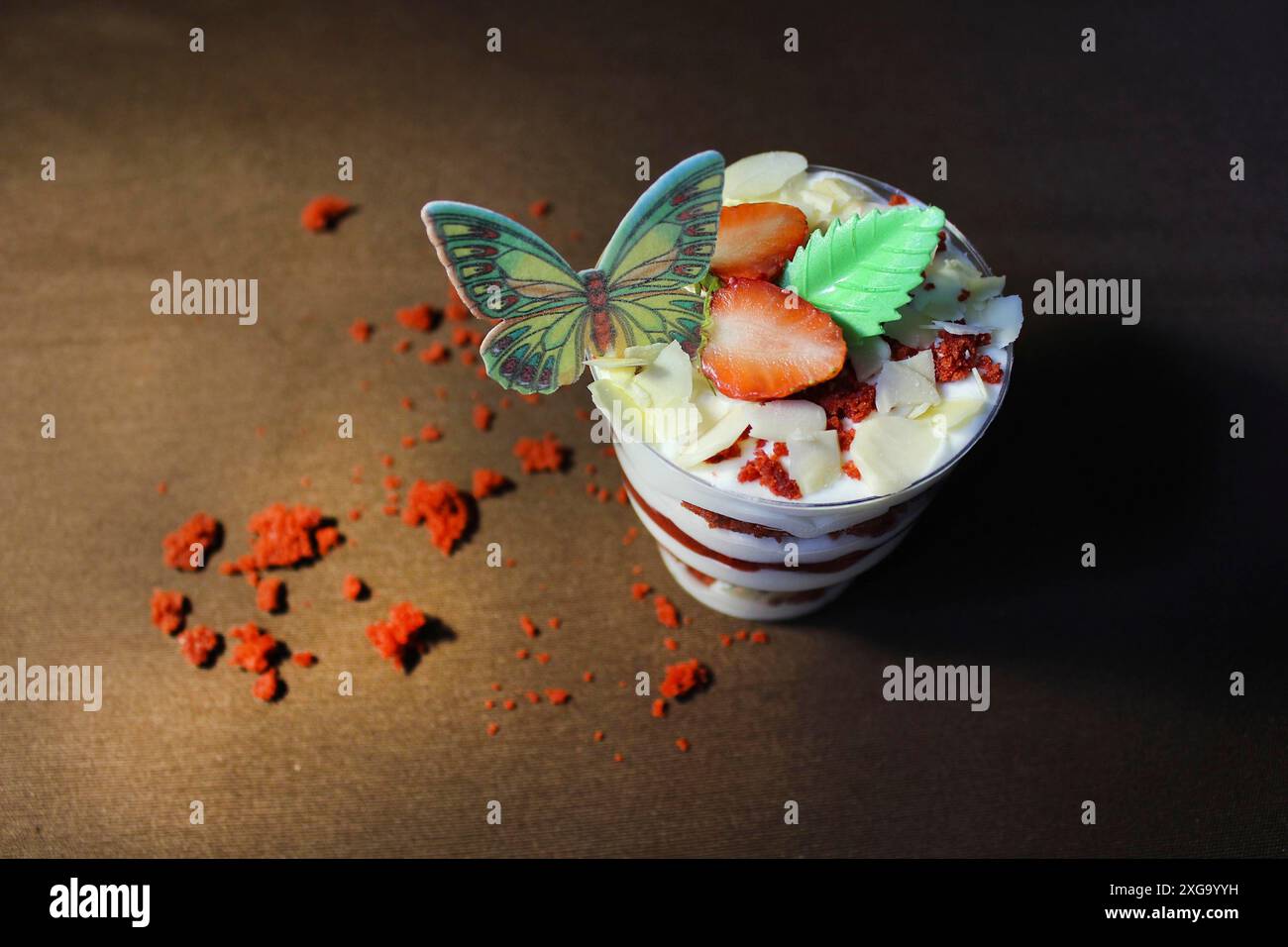 Mehrschichtiges Dessert mit rotem Samtkuchen, Erdbeeren, Creme und dekorativem Schmetterling und Blatt auf Holzuntergrund Stockfoto