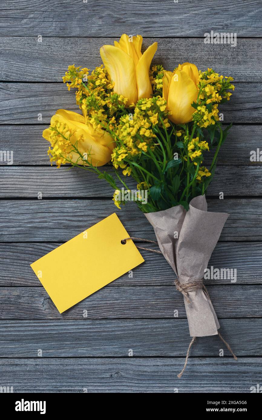 Strauß von gelben Blumen mit leerer Papierkarte als Tag für Namen oder Grüße auf Holzhintergrund Stockfoto