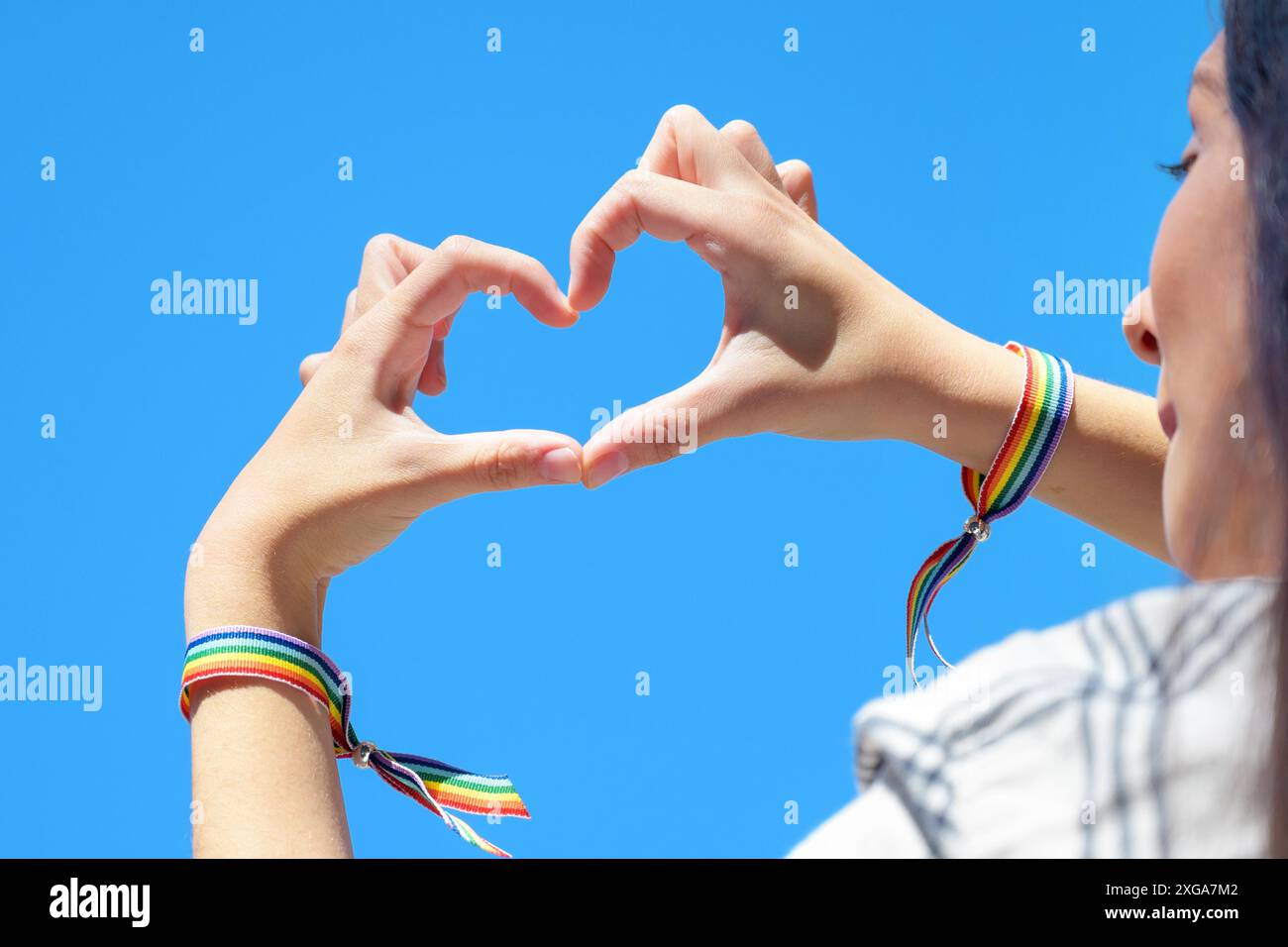 Nahaufnahme von Frauenhänden in einem Regenbogenarmband, die eine Herzform auf blauem Himmel bilden. Hochwertige Fotos Stockfoto