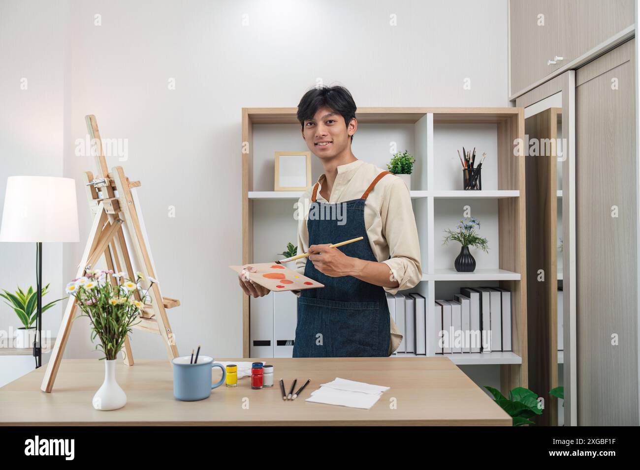 Junger Künstler malte im modernen Studio mit Leinwand und Kunstbedarf, lächelnd in die Kamera, umgeben von Pflanzen und Bücherregalen Stockfoto