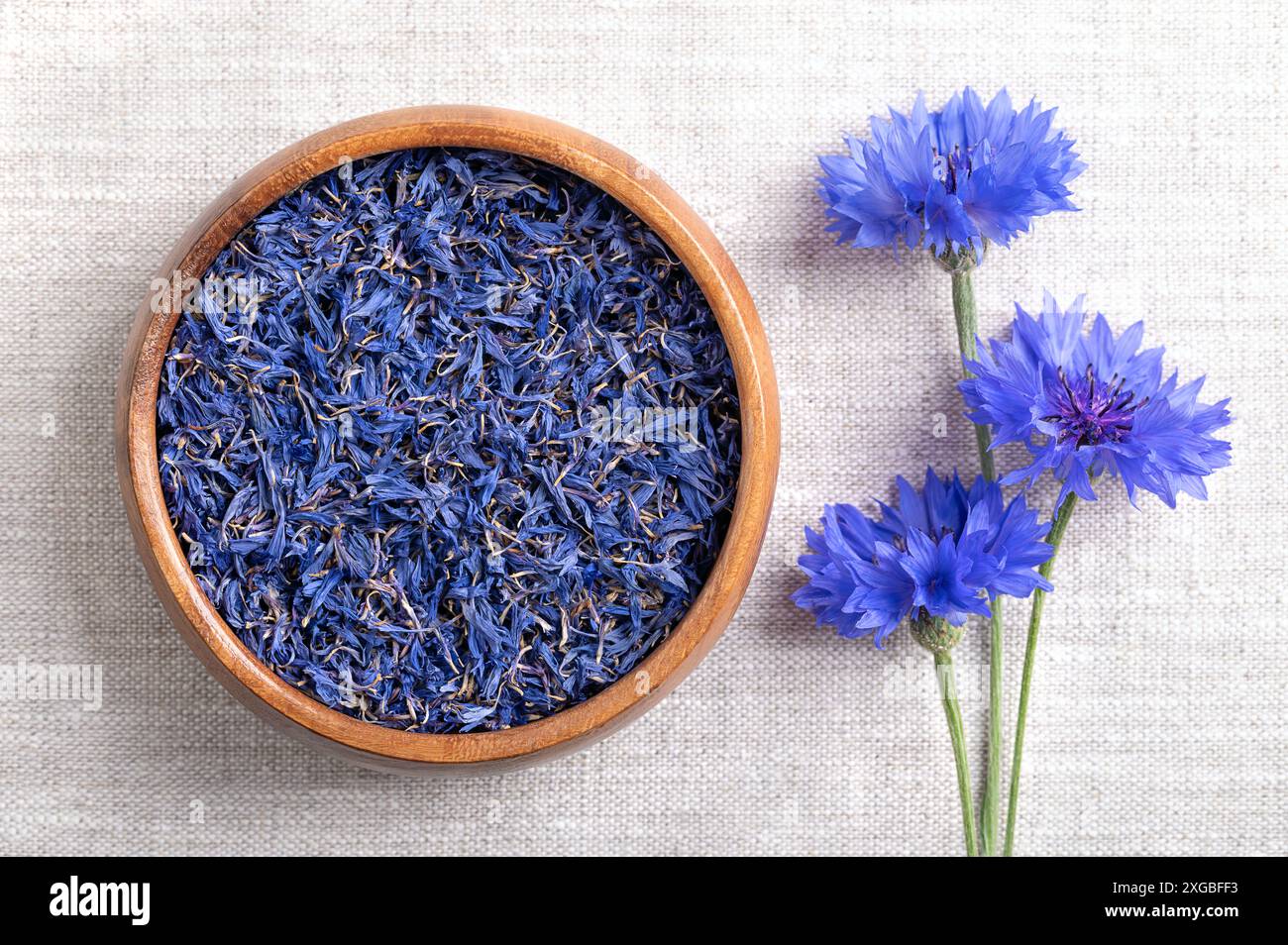 Getrocknete blaue Kornblumenblätter in Holzschale auf Leinengewebe. Essbare Blüten von Centaurea cyanus, auch bekannt als Junggeselenknopf. Stockfoto