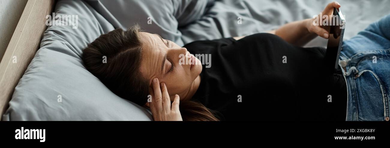 Frau mittleren Alters, die im Bett liegt und eine Tablette mit nachdenklichem Ausdruck hält. Stockfoto