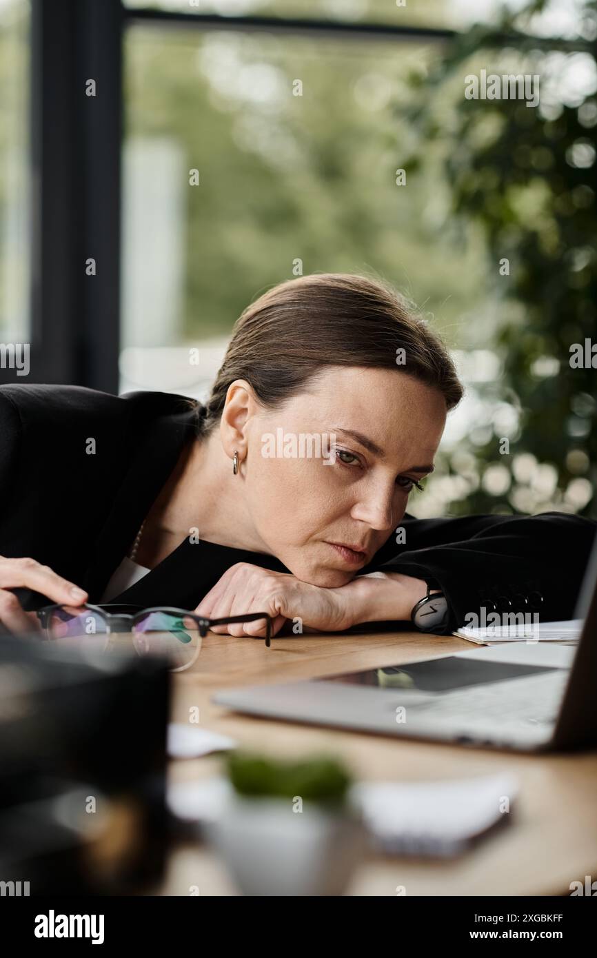 Eine Frau mittleren Alters, die Stress erlebt, wird in einem Büro über ihren Laptop gebeugt. Stockfoto