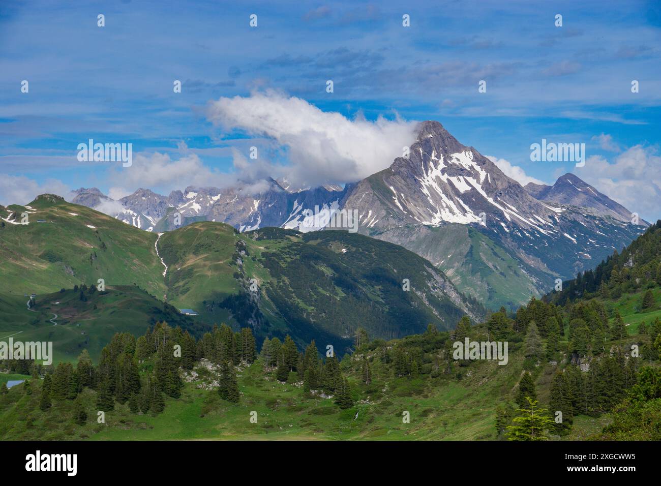 Panorama mit Parkbank auf einem Wanderweg mit Blick auf einen See und die umliegenden Berge im Arlberg, schneebedeckter Wolkengipfel Stockfoto