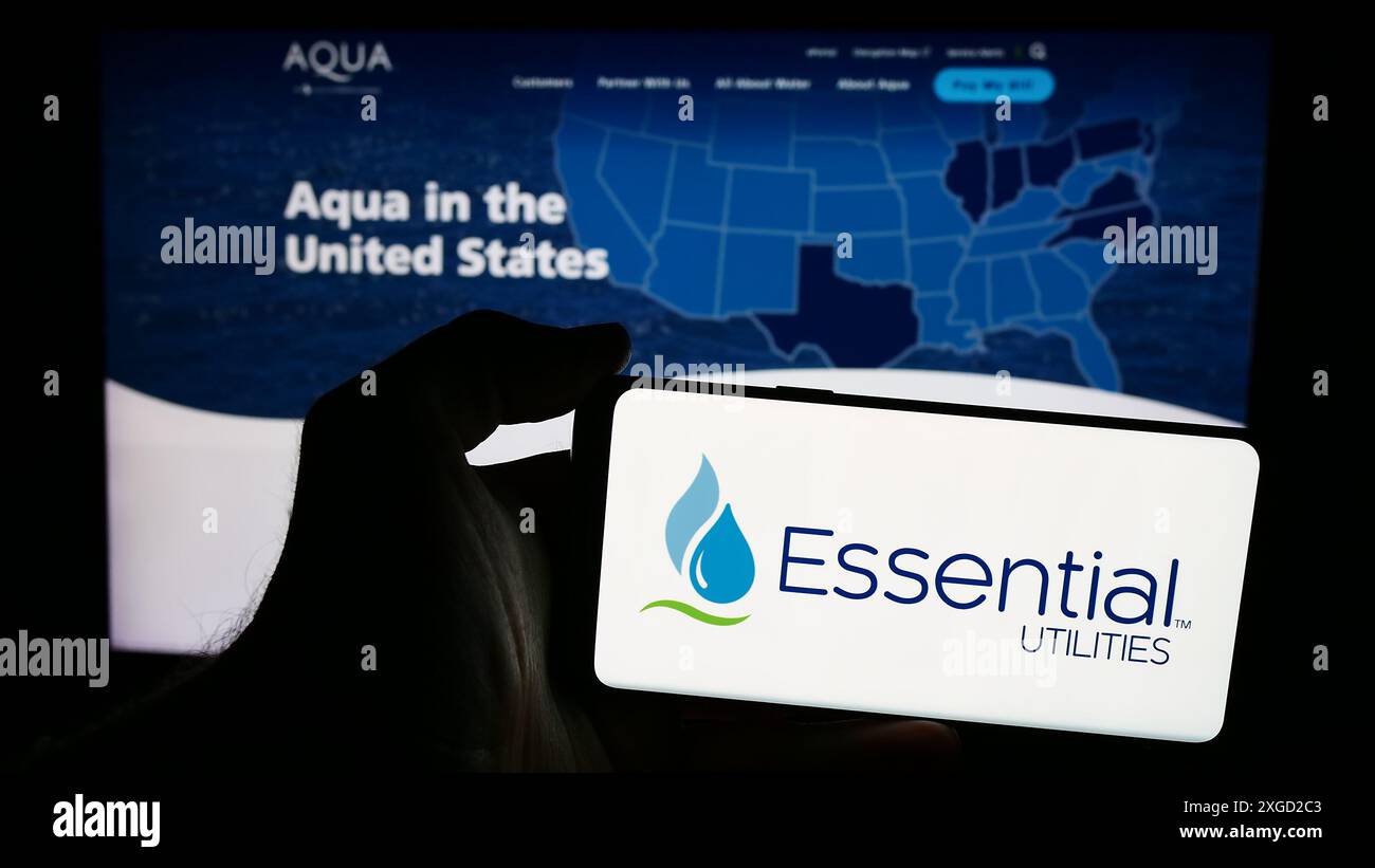 Person mit Smartphone und Logo des US-Wasserversorgungsunternehmens Essential Utilities Inc. Vor der Website. Schwerpunkt auf dem Display des Telefons. Stockfoto