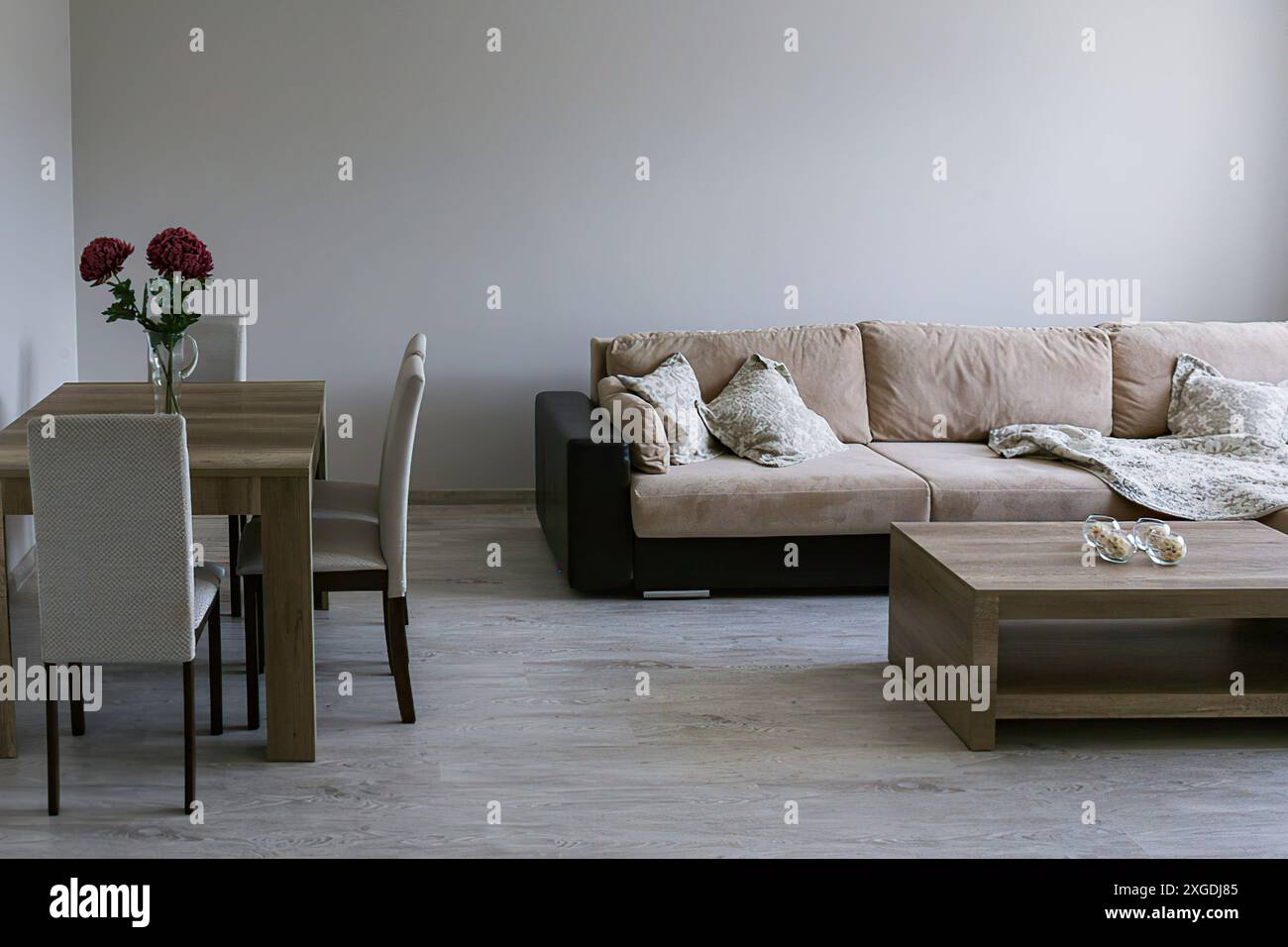 Ein Sofa mit Überwürfen und Kissen, ein Couchtisch, ein Tisch mit drei Stühlen, eine Vase mit roten Blumen, ein hellgrauer Boden und Wände. Stockfoto