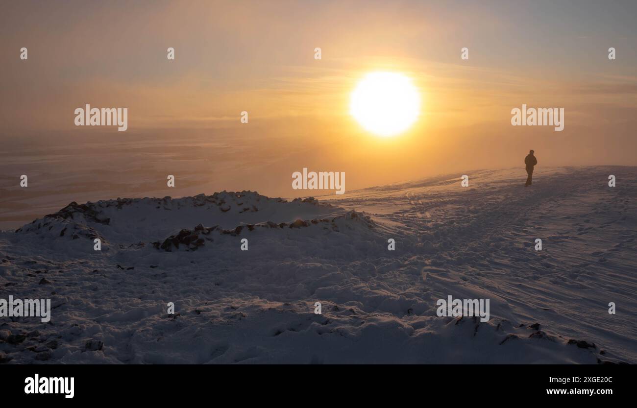 Person, die in Richtung der untergehenden Sonne auf einem schneebedeckten Berggipfel in den Pentlands Hills in der Nähe von Edinburgh, Schottland, geht. Die Sonne untergeht im Hintergrund. Stockfoto