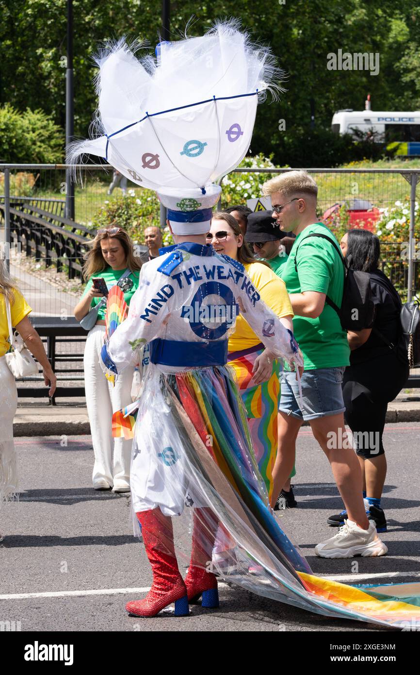 Eine Person, die ein farbenfrohes Kostüm mit einem ausgeklügelten Kopfschmuck, Stiefeln und einem Statement „Mind the Weather“ trägt, repräsentiert TFL beim London Pride 2024 Stockfoto