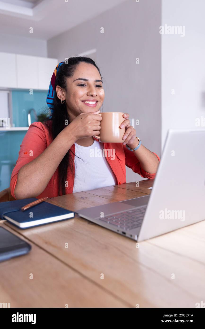 Kaffee trinken und lächeln, Frau, die am Schreibtisch im Heimbüro ihren Laptop benutzt Stockfoto