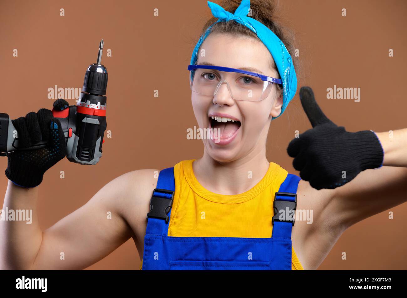 Attraktive junge kaukasische Frau in Arbeitsuniform mit Schutzbrille und einem Schraubenzieher in der Hand, der glücklich lächelt und die Daumen-nach-oben-Geste auf Braun zeigt Stockfoto