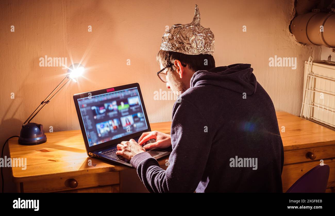 Der junge Mann mit Aluminiumkappe sitzt im dunklen Keller vor einem Laptop. Verschwörungstheorie Konzept Stockfoto