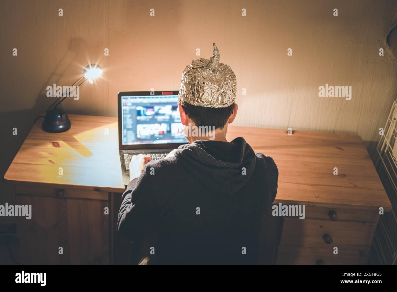 Der junge Mann mit Aluminiumkappe sitzt im dunklen Keller vor einem Laptop. Verschwörungstheorie Konzept Stockfoto