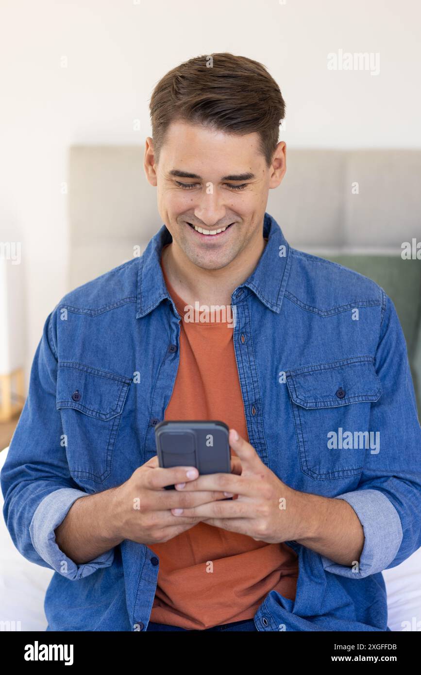 Lächelnder Mann, der das Smartphone benutzt, während er in lässigem Outfit auf dem Bett sitzt Stockfoto