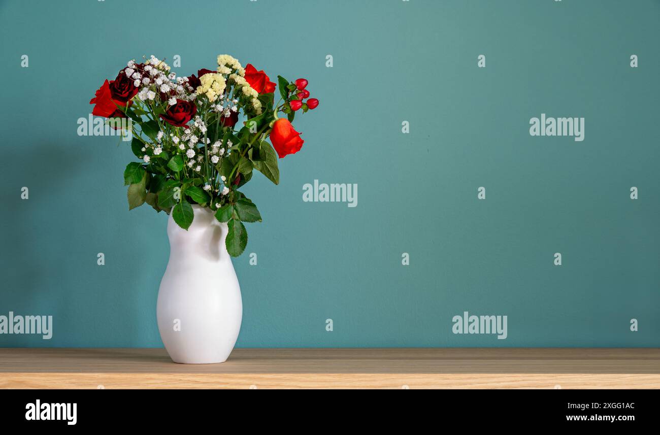 Blumenstrauß aus bunten Rosen in einer weißen Vase auf einem Holztisch vor einer türkisfarbenen Wand mit Kopierraum Stockfoto