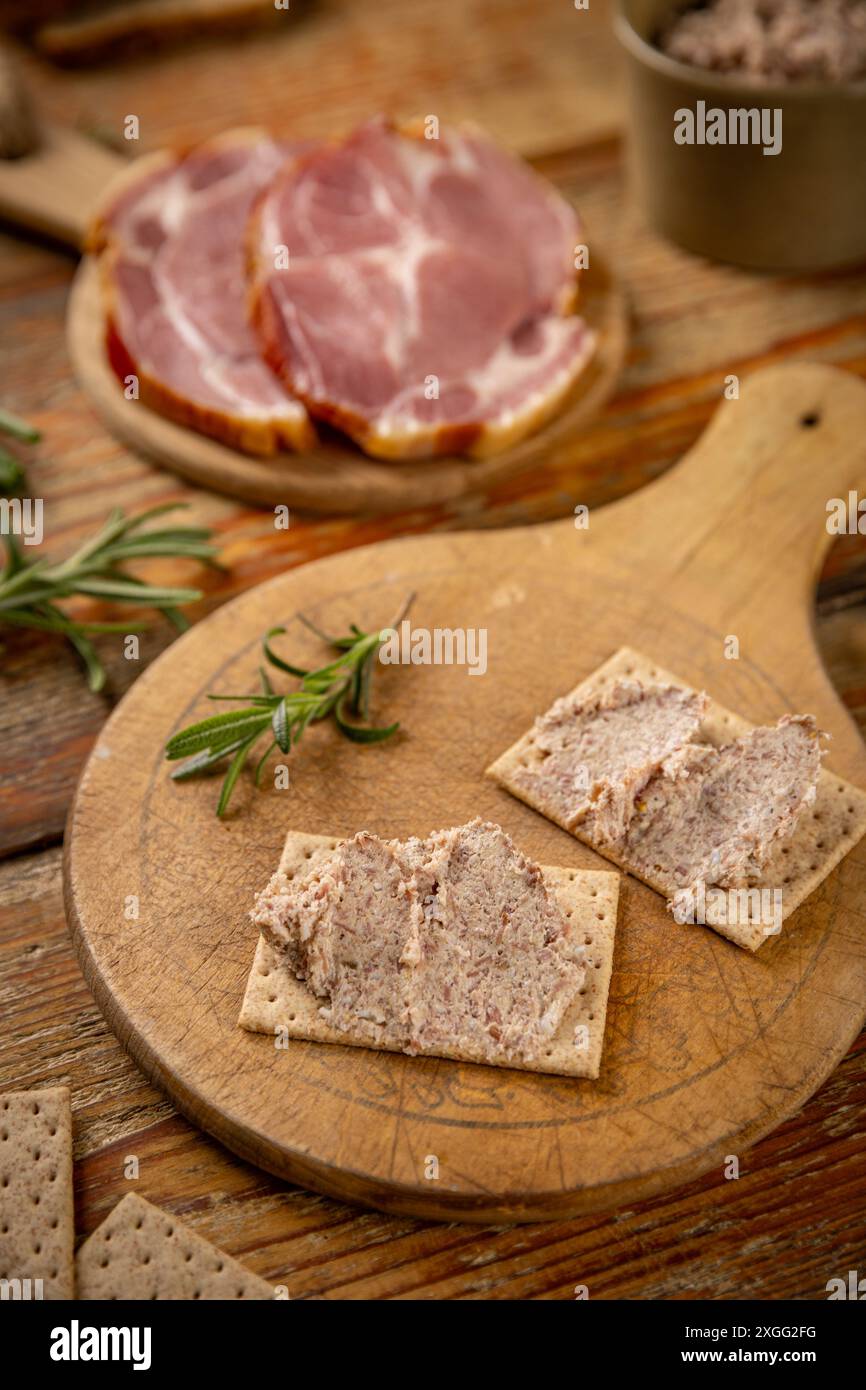 Pastete oder Fleischaufstrich wird auf einem Holzbrett mit frischen grünen Zwiebeln und Rosmarin auf Cracker gelegt Stockfoto