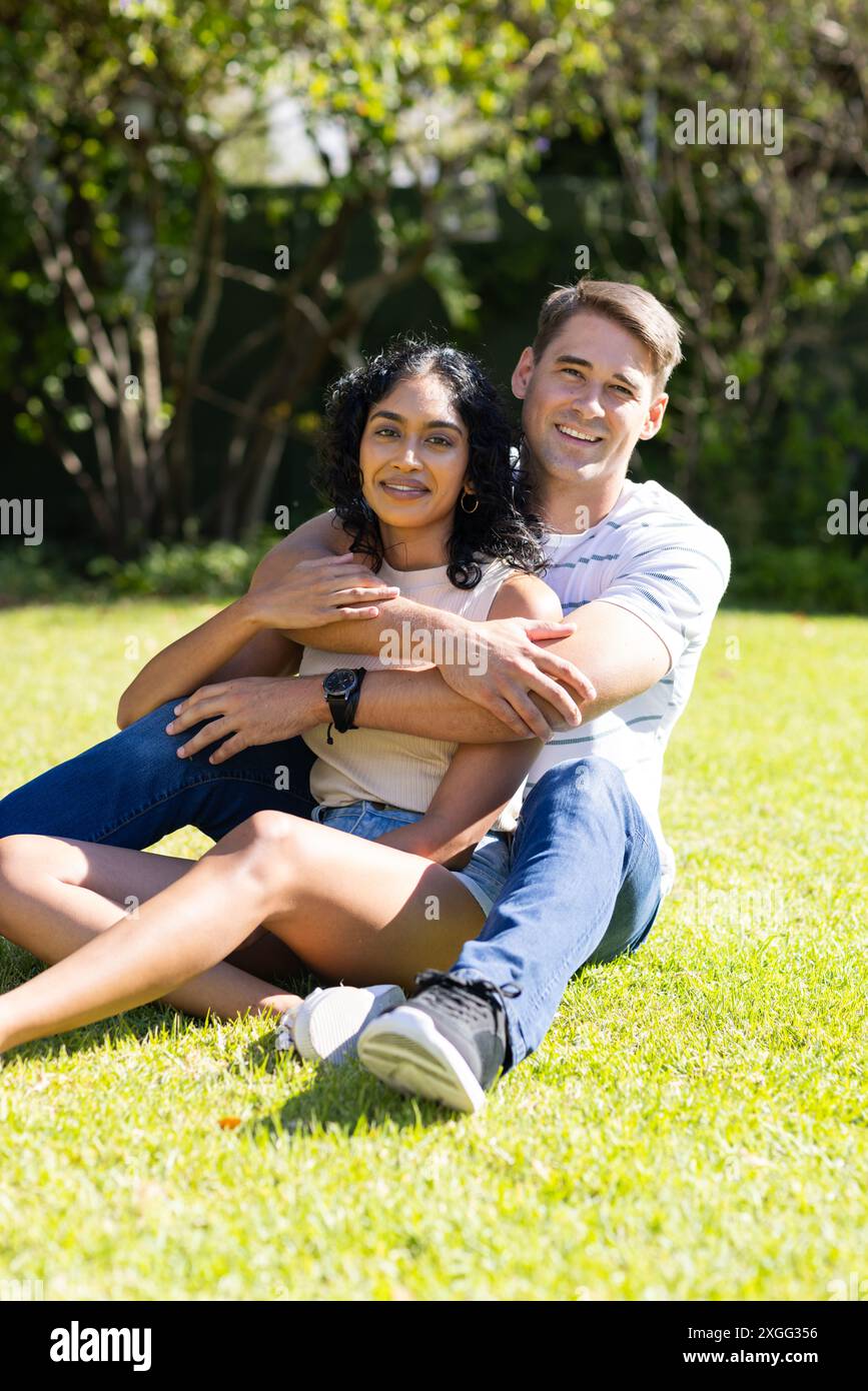 Ein junges Paar, das im Park auf Gras sitzt, umarmt und lächelt in die Kamera Stockfoto