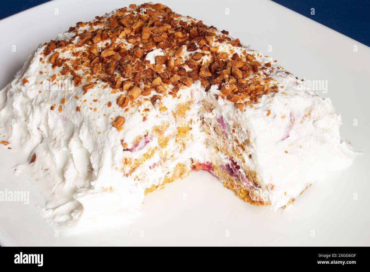 Nahaufnahme eines hausgemachten Desserts mit einer herausgenommenen Scheibe, garniert mit Schlagsahne und bestreut mit knusprigen Nussstückchen, serviert auf einer makellos weißen pla Stockfoto