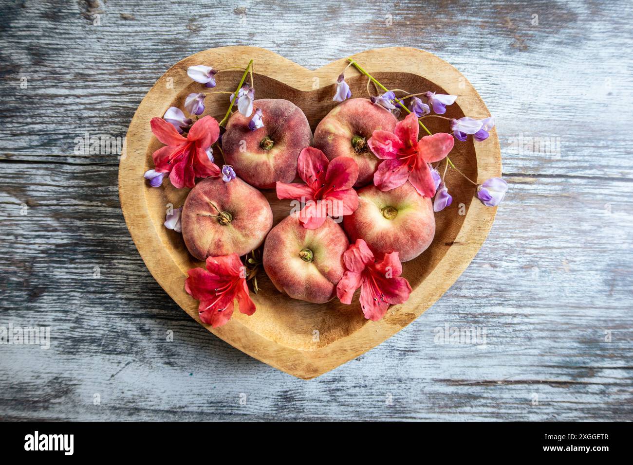 Pfirsiche in einer herzförmigen Holzschale auf einem Tisch mit verstreuten Sommerblumen Stockfoto