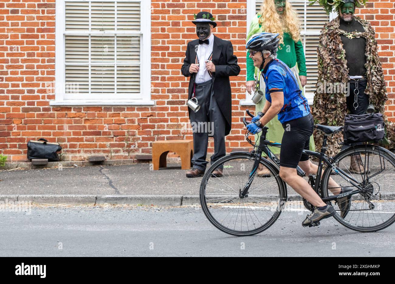 Im Dorf Rendham in Suffolk markieren die Mummers auf dem Bürgersteig die Sommersonnenwende mit einer Show mit einem unwahrscheinlichen Passanten auf einem Fahrrad auf der Straße. Stockfoto