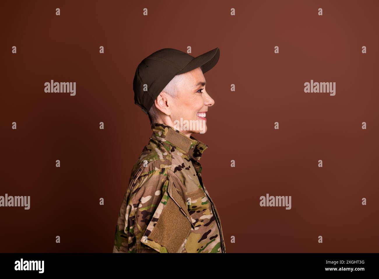 Foto von attraktiver positiver positiver älterer Dame mit Camo-Uniform, die leeren Raum aussieht, isolierter brauner Hintergrund Stockfoto