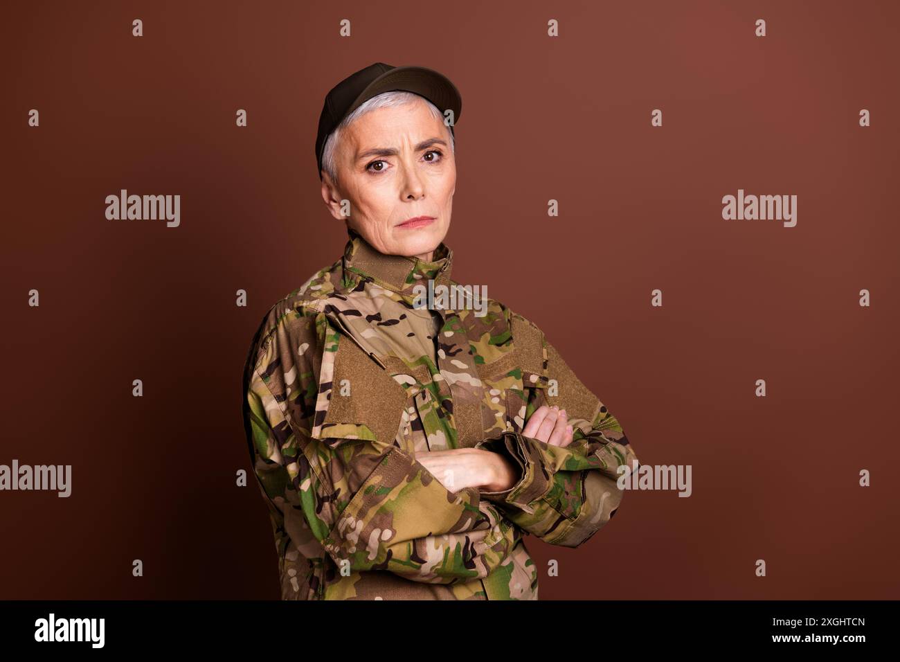 Foto von ernster selbstbewusster älterer Dame gekleidet Camo Uniform Arme gefaltet leeren Raum isoliert brauner Hintergrund Stockfoto