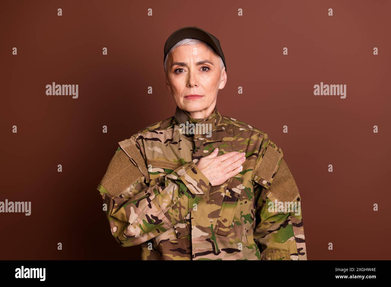 Foto von ernster zuversichtlicher älterer Dame gekleidet Camo Uniform hören Hymne Arm Hand Brust leerer Raum isolierter brauner Hintergrund Stockfoto