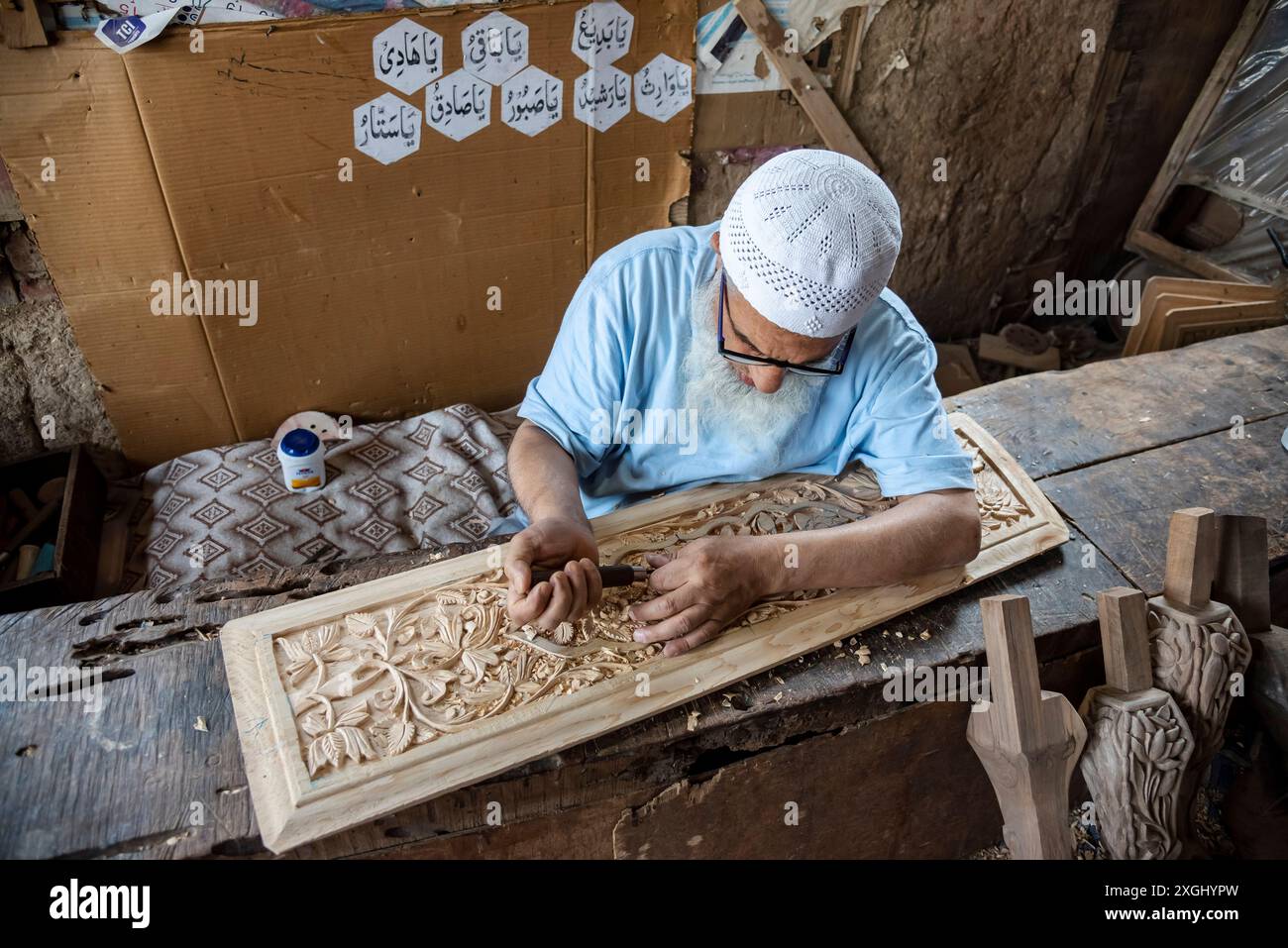 In seiner Werkstatt in Srinagar wird ein Meister gesehen, wie er Entwürfe an einem Holzstück herstellt. Kaschmir ist bekannt für seine exquisite Holzschnitzerei, ein Handwerk, das über Generationen weitergegeben wurde. Aus strapazierfähigem Walnussholz kreieren erfahrene Handwerker komplizierte Blumenmuster, geometrische Designs und traditionelle Motive. Diese akribische Arbeit, die auf Möbeln, dekorativen Tafeln und Souvenirs zu sehen ist, zeigt das kulturelle Erbe der Region. Ghulam Nabi Zargar, ist ein Meisterschnitzer, der detaillierte Entwürfe auf eine Walnussholz-Holzoberfläche graviert. Das Licht hebt die komplizierten Muster hervor und erweckt die Kunst zum Leben Stockfoto