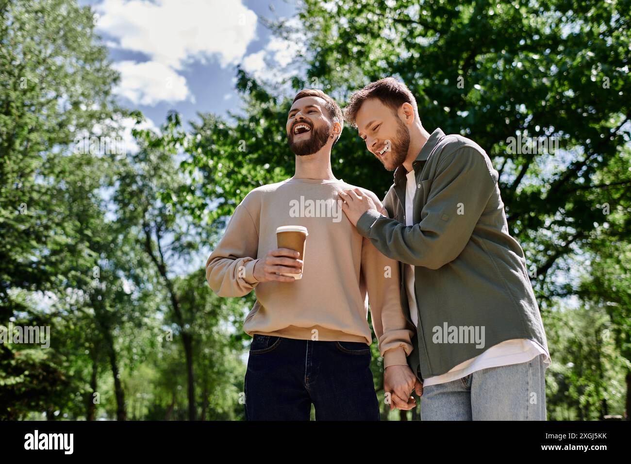 Ein bärtiges Schwulenpaar lacht an einem sonnigen Tag durch einen Park. Stockfoto