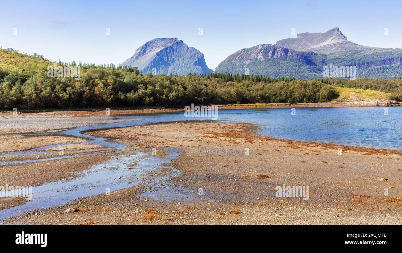 Ruhiger Strand mit einem sich windenden Bach und majestätischen Bergen unter einem klaren blauen Himmel entlang der malerischen Nationalstraße Helgelandkysten. Nordnorwegen Stockfoto