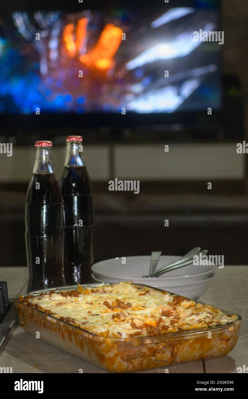 Gönnen Sie sich eine hausgemachte Lasagne, die heiß und frisch aus dem Ofen serviert wird, begleitet von zwei Flaschen Cola. Perfekt für einen gemütlichen Filmabend zu Hause. Diese Inv Stockfoto