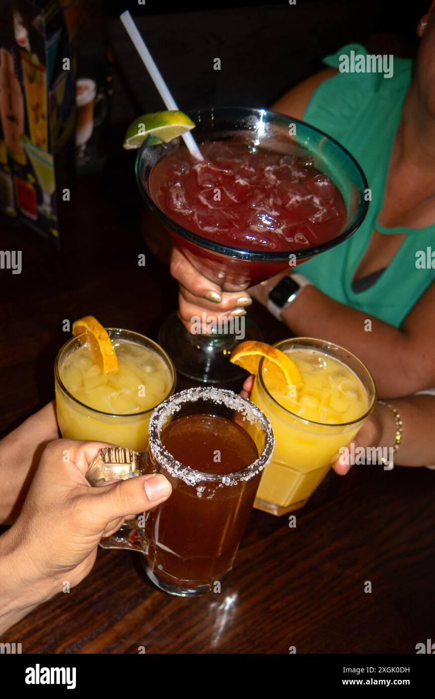 Lebhaftes Bild von Freunden, die mit einer Vielzahl von Cocktails, darunter eine große Margarita, zwei Orangengetränke und ein Bier mit gesalzenem Rand, toasten. Celebrato Stockfoto