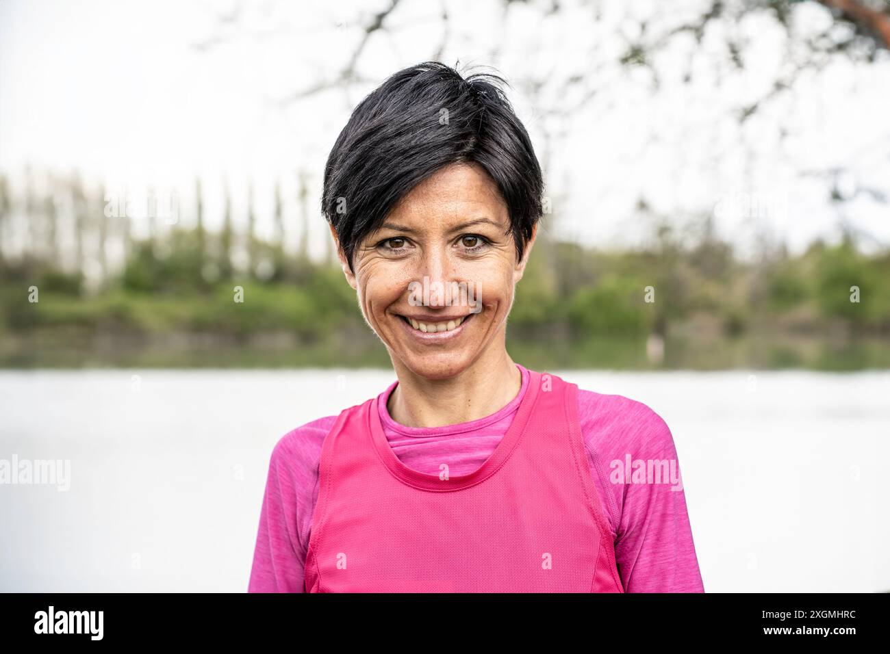 Porträt einer lächelnden Frau mittleren Alters mit kurzen schwarzen Haaren in rosa Sportkleidung, die draußen am See steht. Konzept des aktiven Lebensstils und Stockfoto