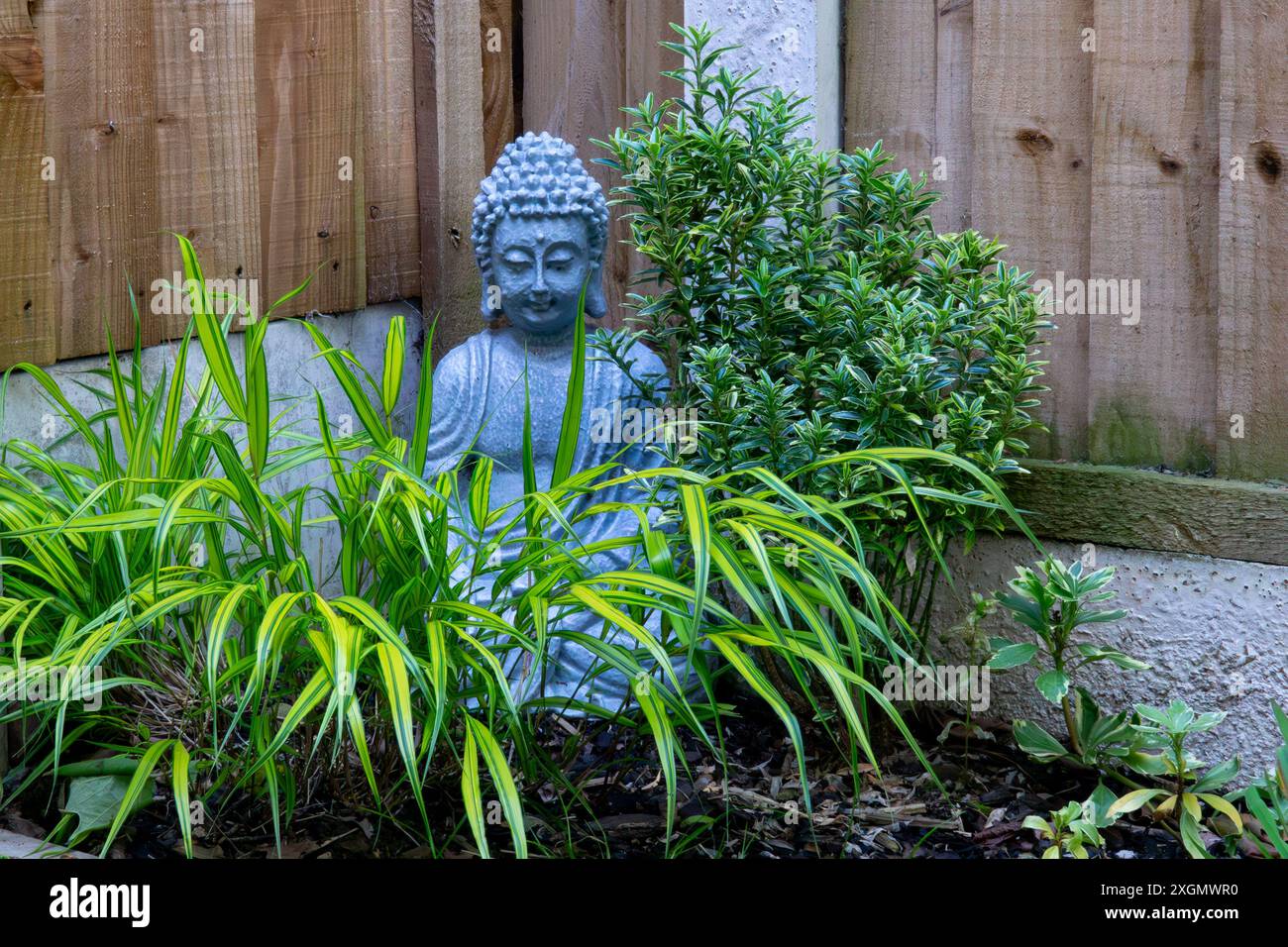 Eine graue Buddha-Statue aus Stein befindet sich in einer ruhigen Gartenumgebung, umgeben von üppig grünem Laub. Stockfoto