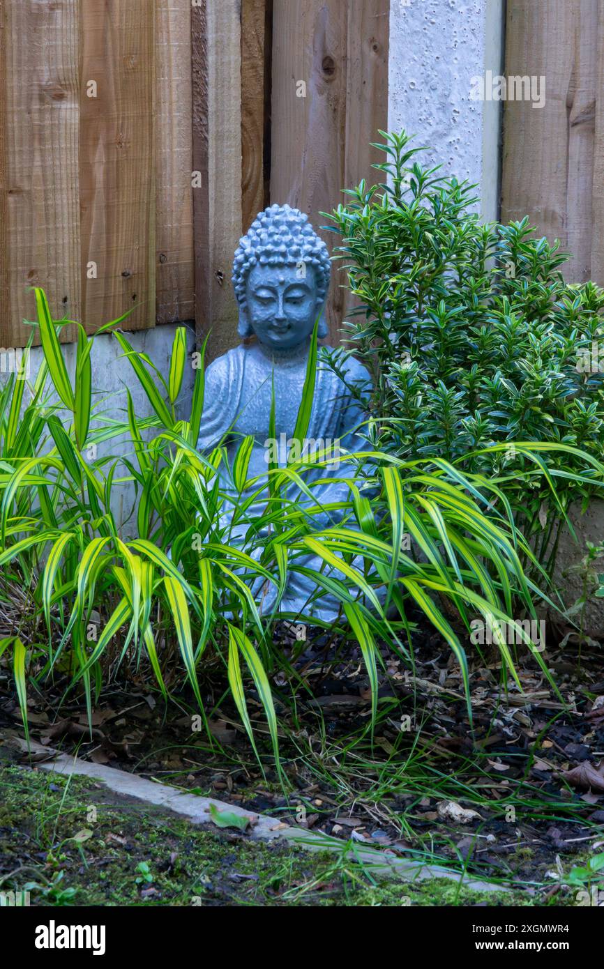 Eine graue Buddha-Statue aus Stein befindet sich in einer ruhigen Gartenumgebung, umgeben von üppig grünem Laub. Stockfoto