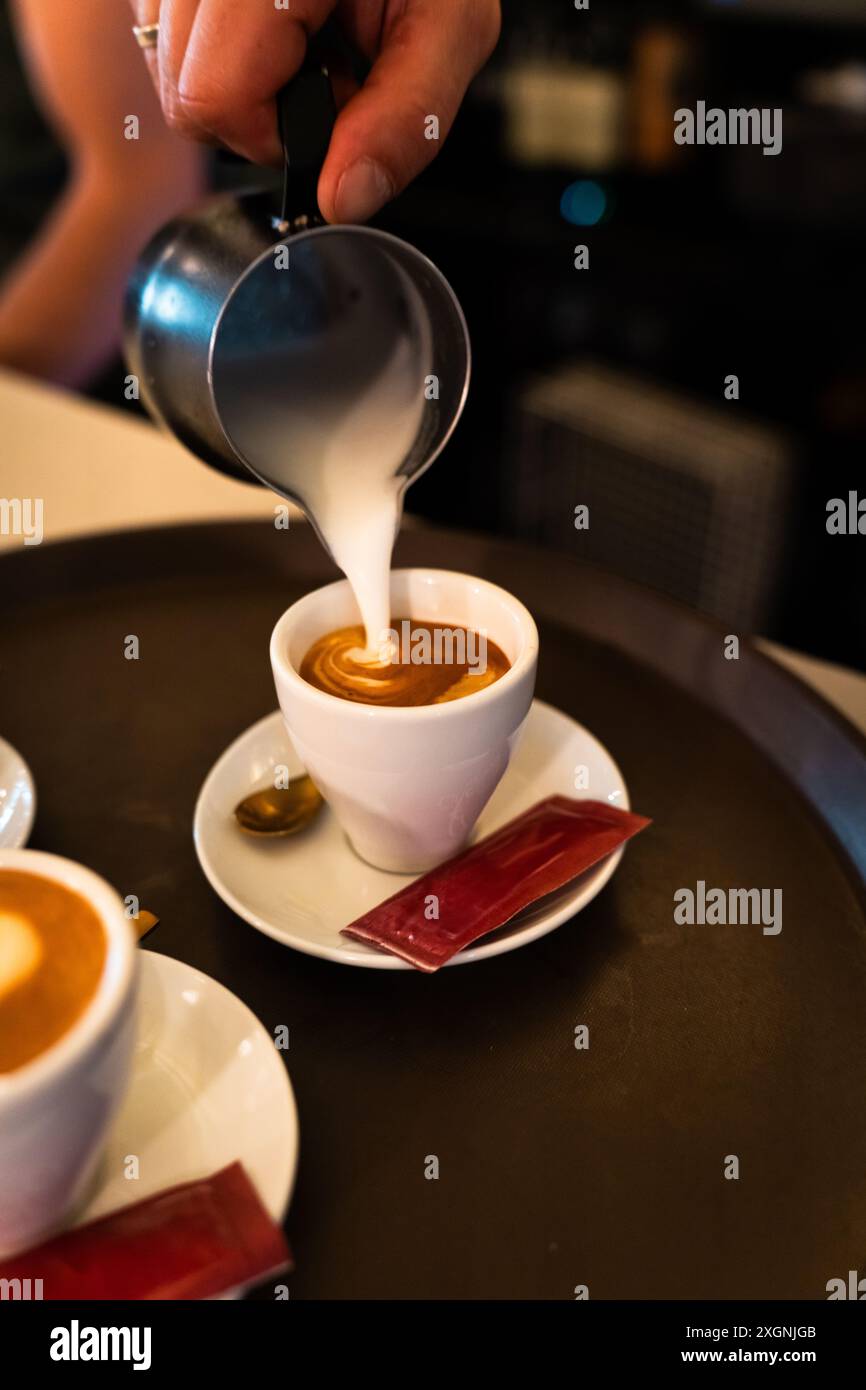 Die Hand des Kellners gießt Milch mit dem Krug in eine Kaffeetasse. Vertikal Stockfoto