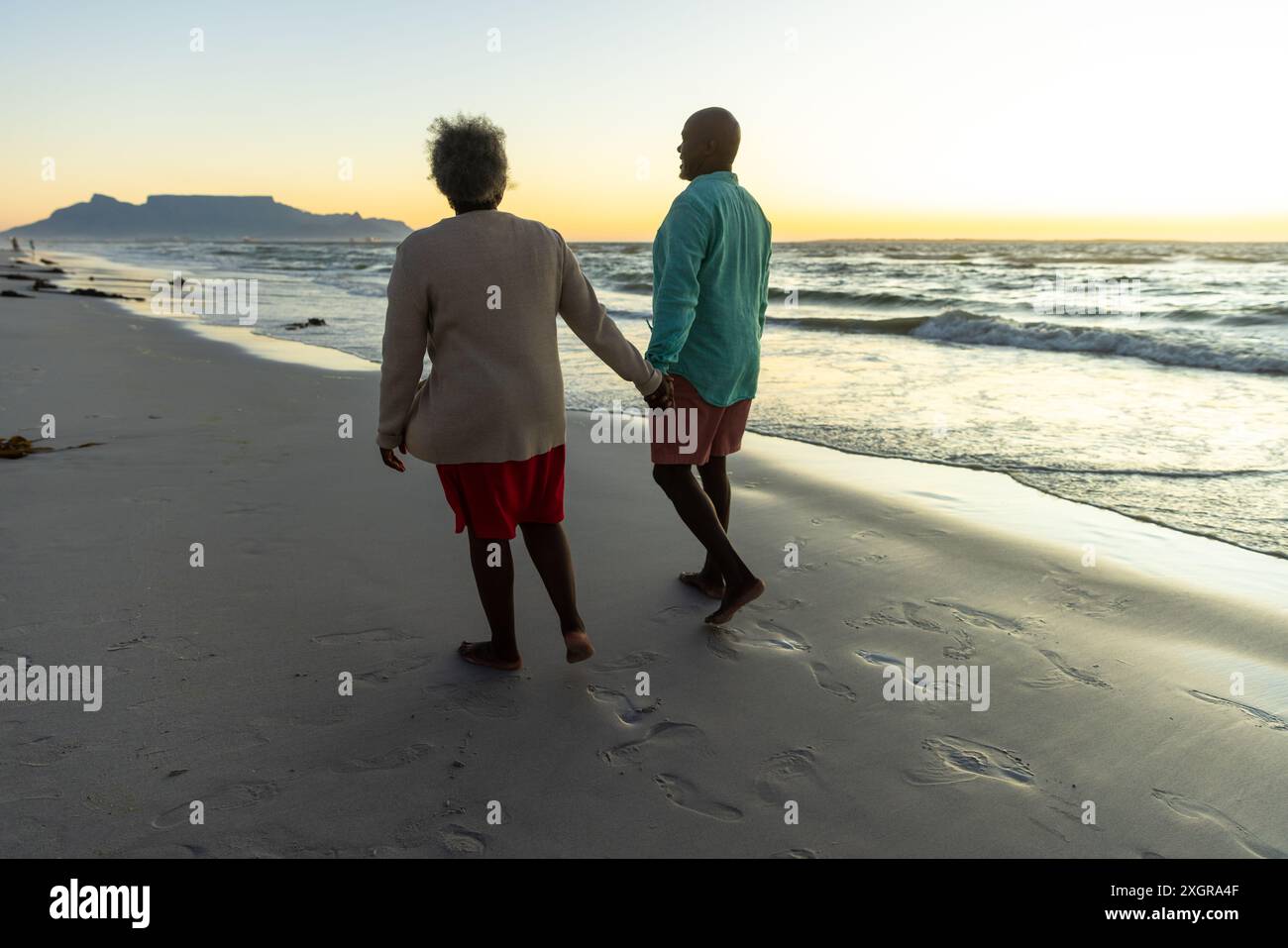 Senior afroamerikanisches Paar genießt einen Strandspaziergang bei Sonnenuntergang. Ihr ruhiger Spaziergang entlang der Küste markiert einen Moment friedlicher Gesellschaft. Stockfoto