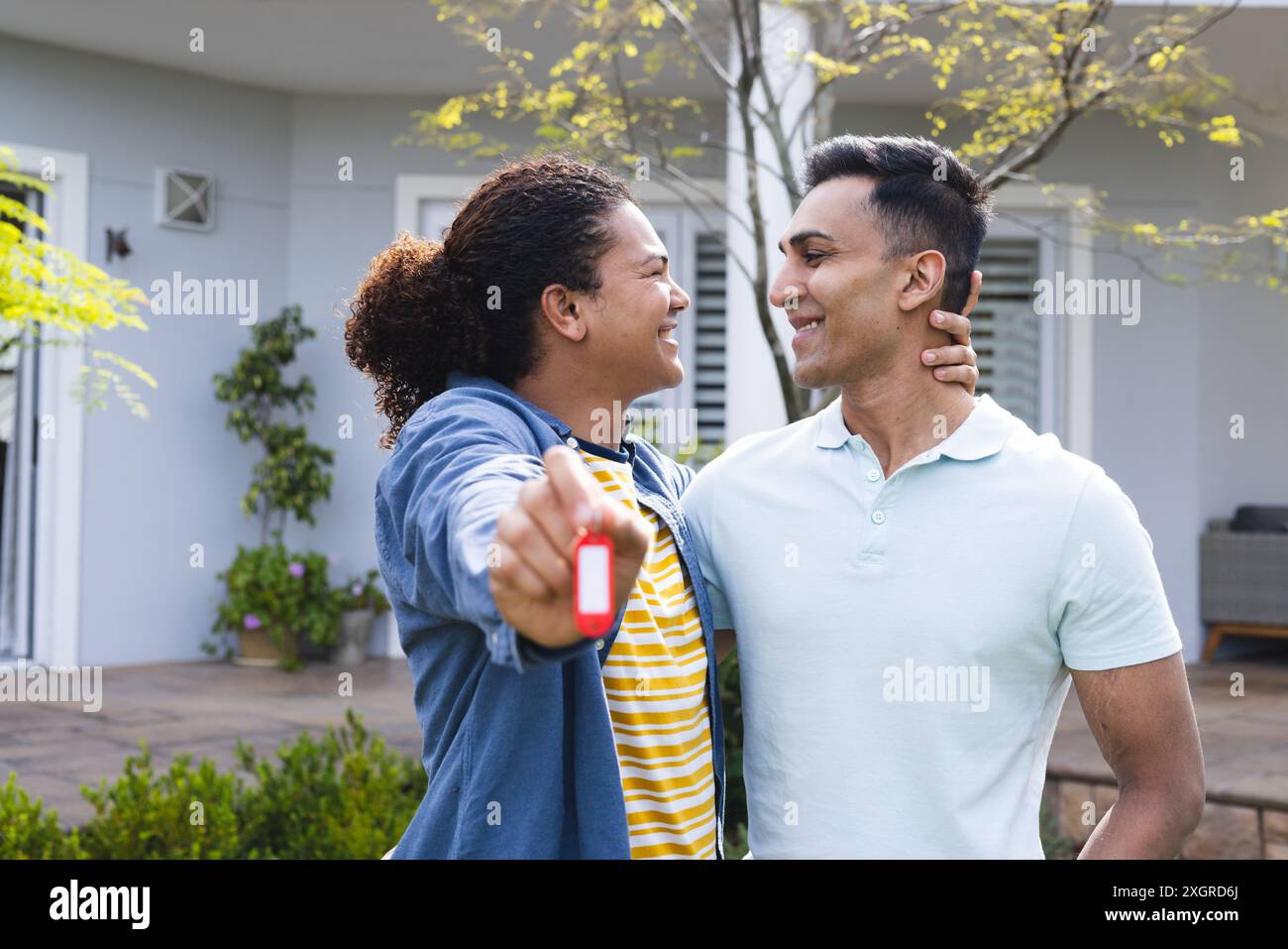 Junge Menschen mit Rasse und nahöstlicher Herkunft lächeln draußen und halten Schlüssel. Sie feiern Hausbesitz in einer sonnigen Vorstadt-Umgebung. Stockfoto