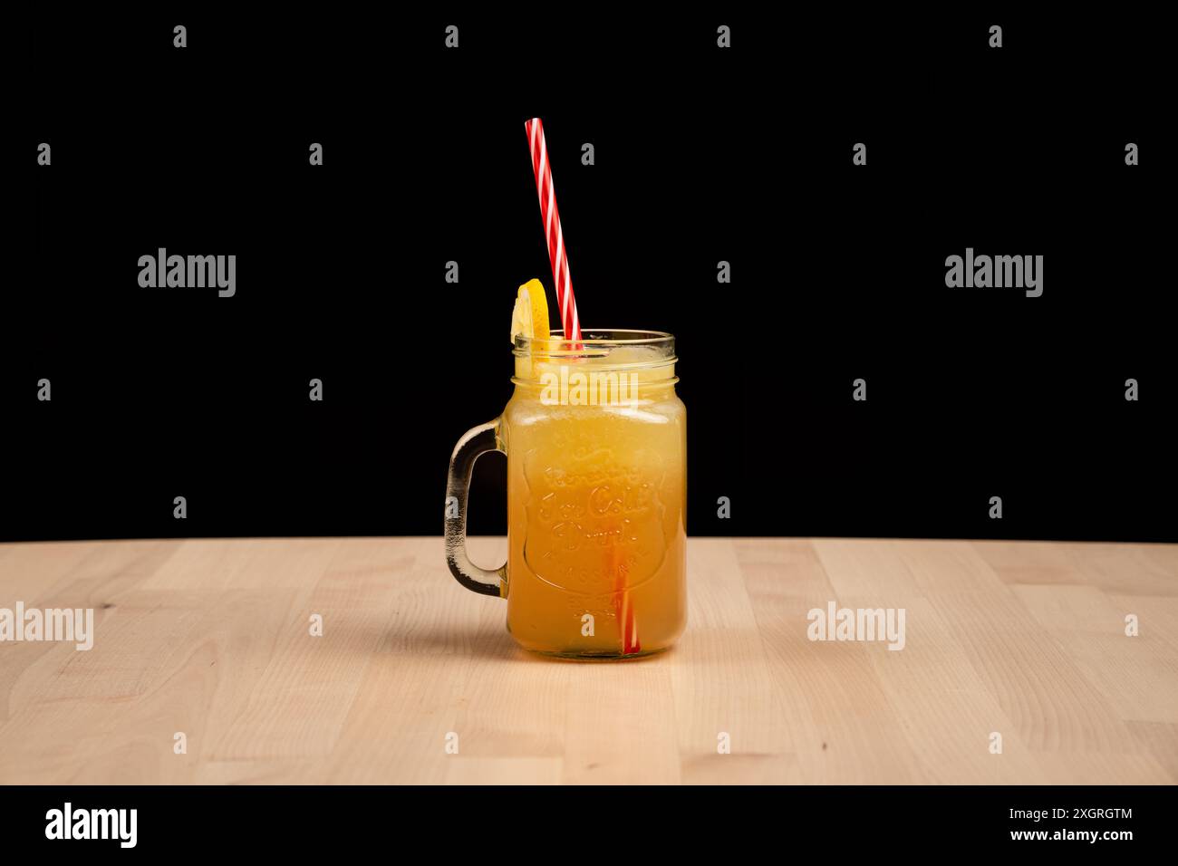 Ein erfrischendes Glas Orangensaft mit rot-weiß gestreiftem Stroh und einer Zitronenscheibe auf Holztisch vor schwarzem Hintergrund Stockfoto