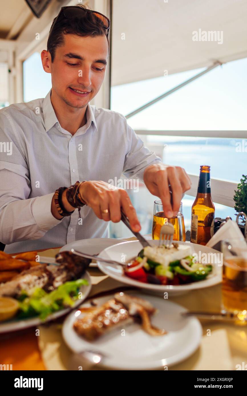 Der Mann isst Fisch mit Gemüse im Restaurant im Freien, serviert mit Getränken. Abendessen mit frischen Meeresfrüchten und Meereslandschaft. Sommerferien Stockfoto Der Mann isst Fisch mit Gemüse im Restaurant im Freien, serviert mit Getränken. Abendessen mit frischen Meeresfrüchten und Meereslandschaft. Sommerferien Stockfoto