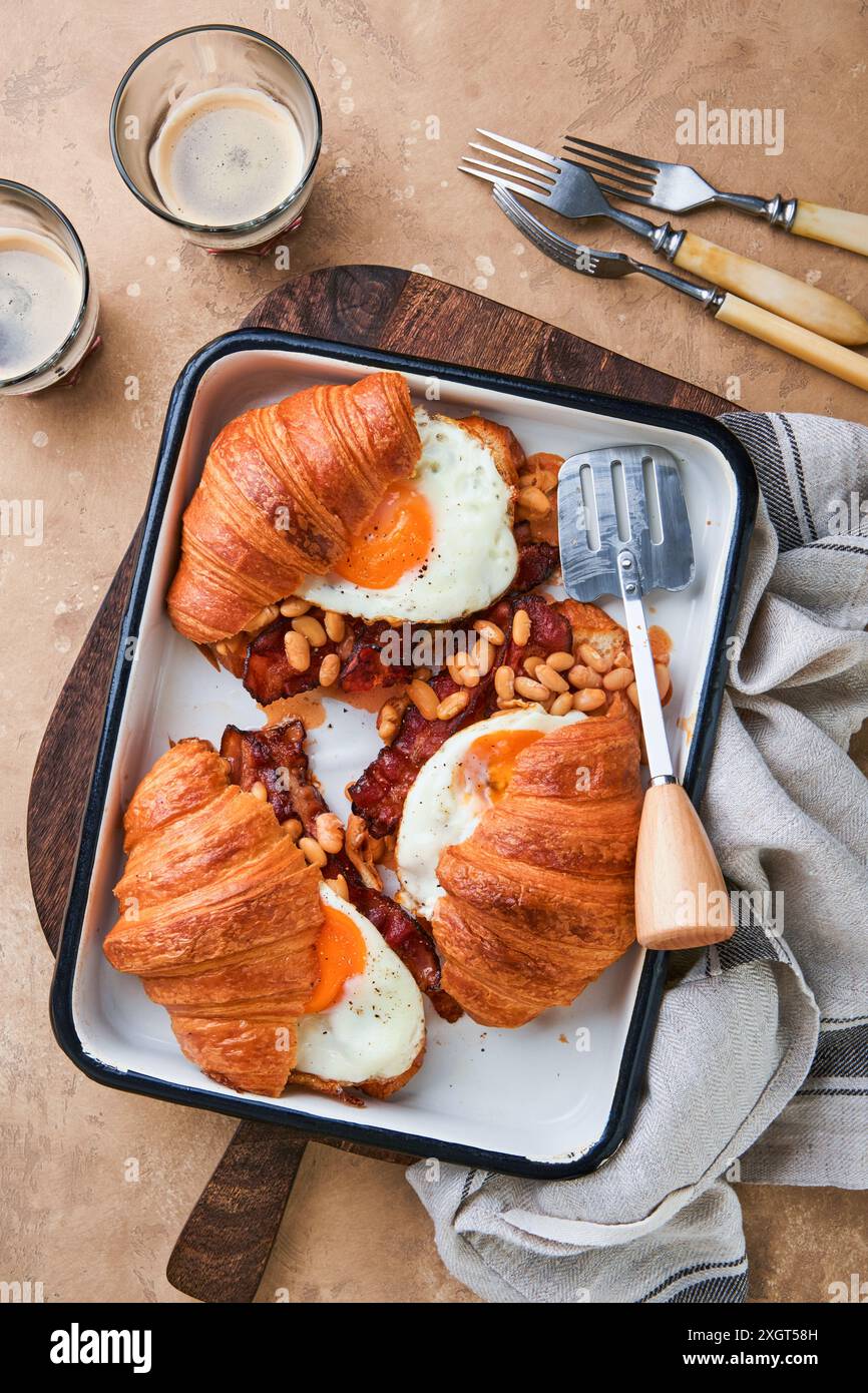 Croissants, englisches Frühstück. Köstliches frisches Frühstück gegrillt drei Croissants mit Speck, Spiegeleiern und Bohnen in Tomatensauce und zwei Gläsern C. Stockfoto